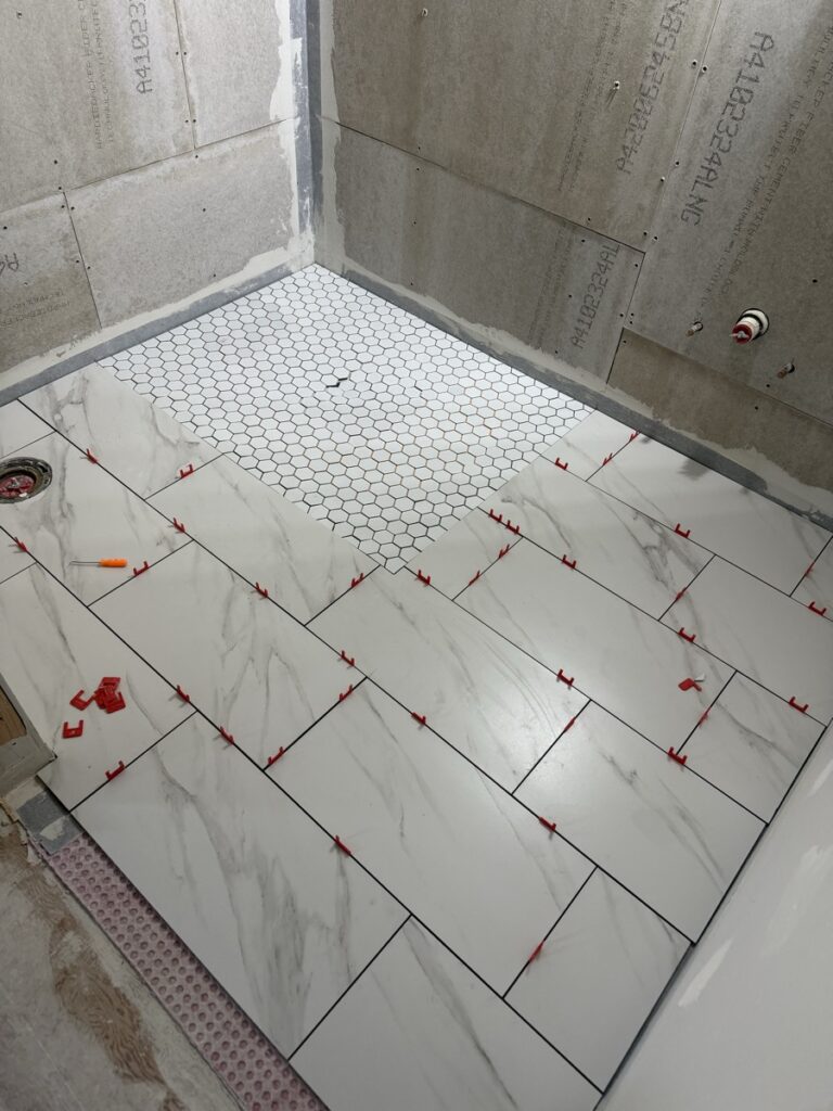Bathroom floor with marble-look porcelain tile and hexagonal mosaic shower pan during installation with leveling clips