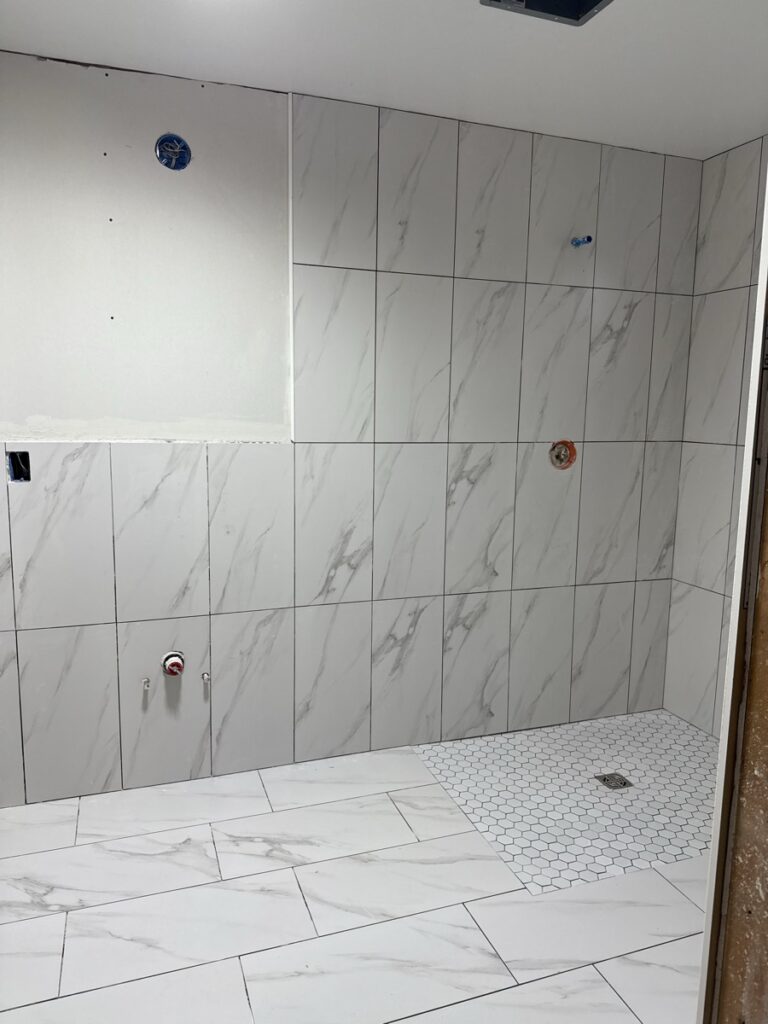 Walk-in shower walls being tiled with marble-look large-format porcelain and mosaic hex shower pan